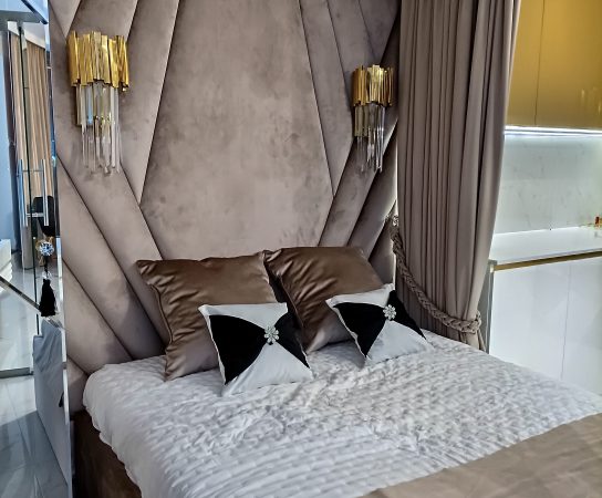 Close-up of the plush, oversized, diamond-tufted velvet headboard in a neutral taupe/beige color, flanked by modern gold and crystal wall sconces.