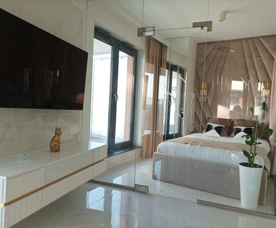 Daytime view of the glass-enclosed bedroom with a large TV mounted on a white and gold console, and floor-to-ceiling windows leading to a balcony.