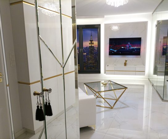 View from the entry/hallway showing mirrored closet doors with gold trim and tassels, reflecting the living area and the skyscraper view outside the window.