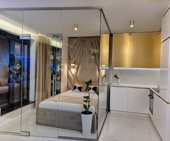 Modern studio apartment bedroom and kitchen separated by a clear glass partition, featuring a plush, tufted velvet headboard and gold kitchen cabinets.