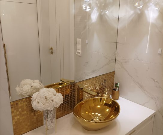 High-end bathroom interior with white marble-effect walls, a gold mosaic backsplash, and a striking golden bowl sink on a white floating vanity.