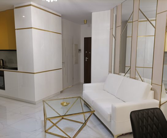 View of the all-white and gold-trimmed kitchen and storage cabinets, and the white sofa and coffee table in the foreground.