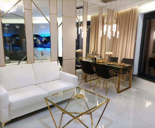 Luxury studio apartment living and dining area in Dubai with white sofa, gold geometric coffee table, black and gold dining set, mirrored wall panels, and crystal chandelier.