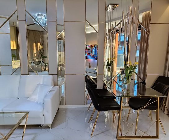 View from the white sofa towards the glass dining table with gold base and black chairs, set against a dramatic, segmented mirrored wall with gold trim.