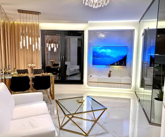Living area view showing a large wall-mounted TV on a marble-effect panel with ambient blue backlighting, a white media console, and a gold-framed glass coffee table.