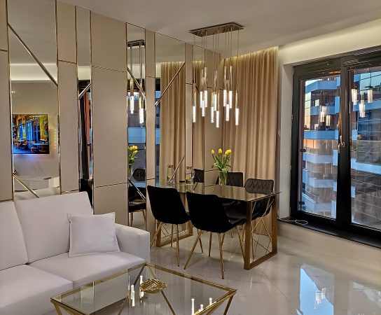 Front view of the white sofa and geometric gold and glass coffee table, with the dining area and mirrored wall visible in the background.